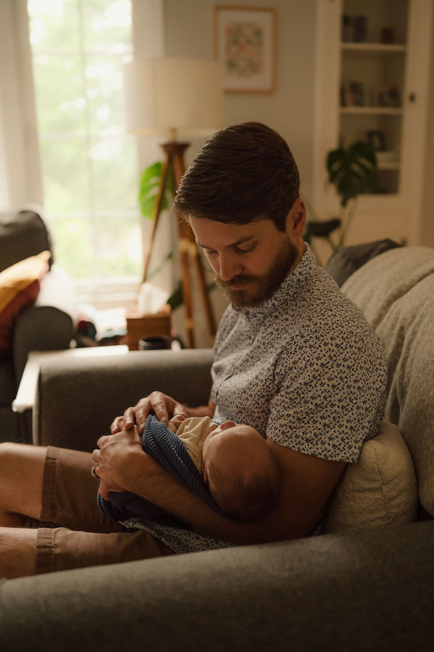 Parent in speckled shirt tenderly holding sleeping infant on dark grey couch in bright room.