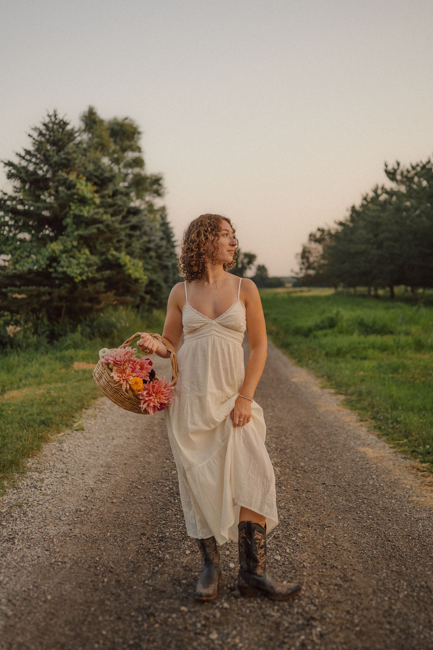 Someone in a light dress and boots walks down a rural path holding summer wildflowers.