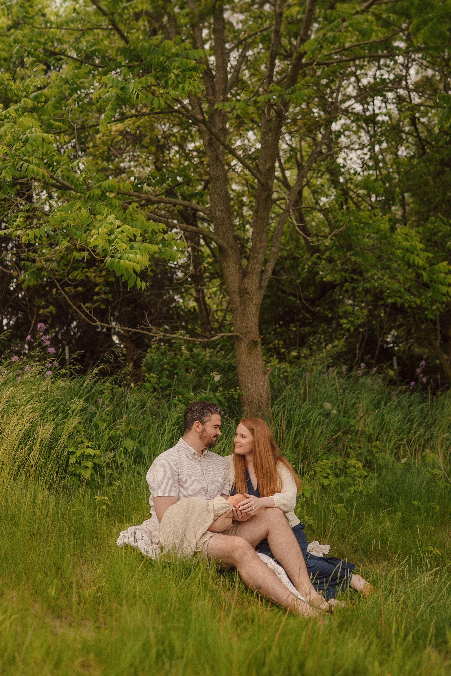 Couple sitting intimately under green tree in summer meadow.