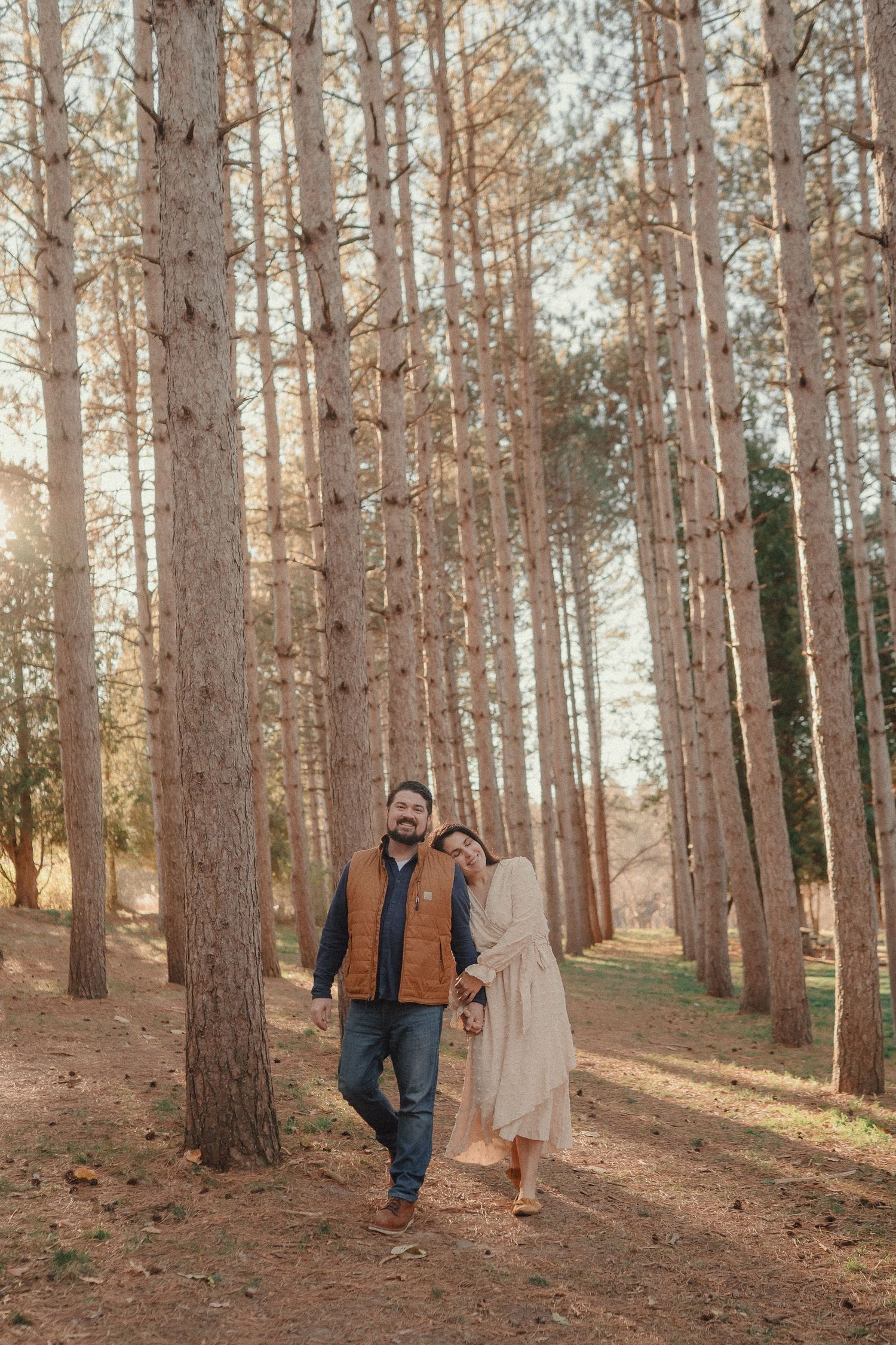 A couple walks through a sunlit pine forest holding hands on a golden autumn afternoon.