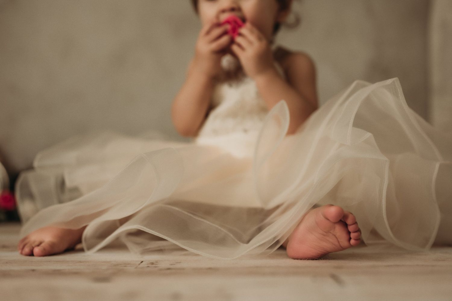 Little bare feet peek out from layers of cream tulle fabric on wooden flooring.