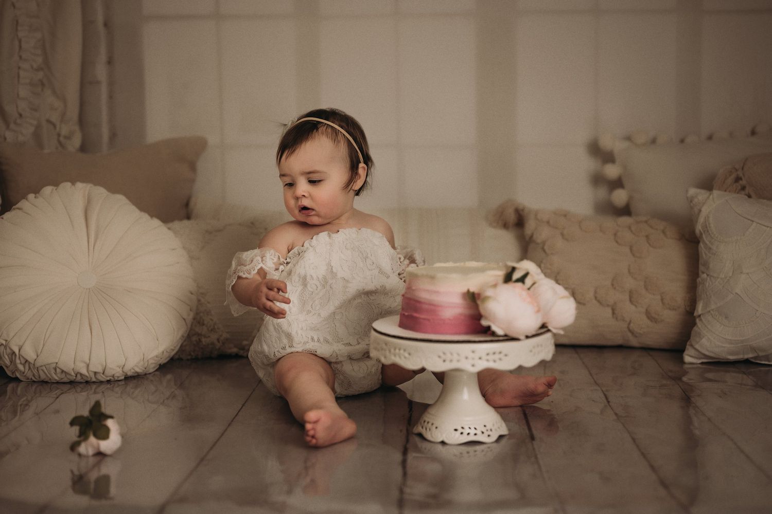 Baby in white lace romper sitting next to pink cake on decorative stand with round pillows in background.