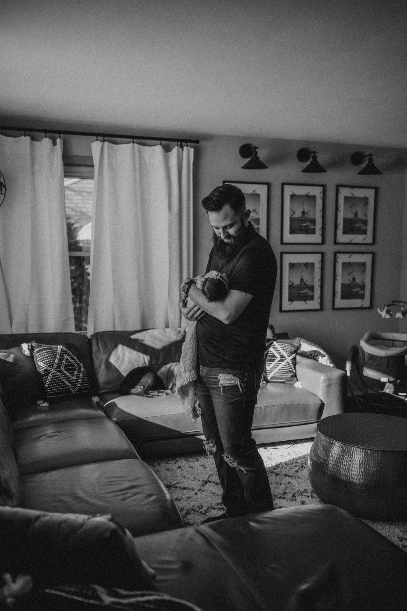 Black and white photo of a parent holding baby in living room with framed wall art.