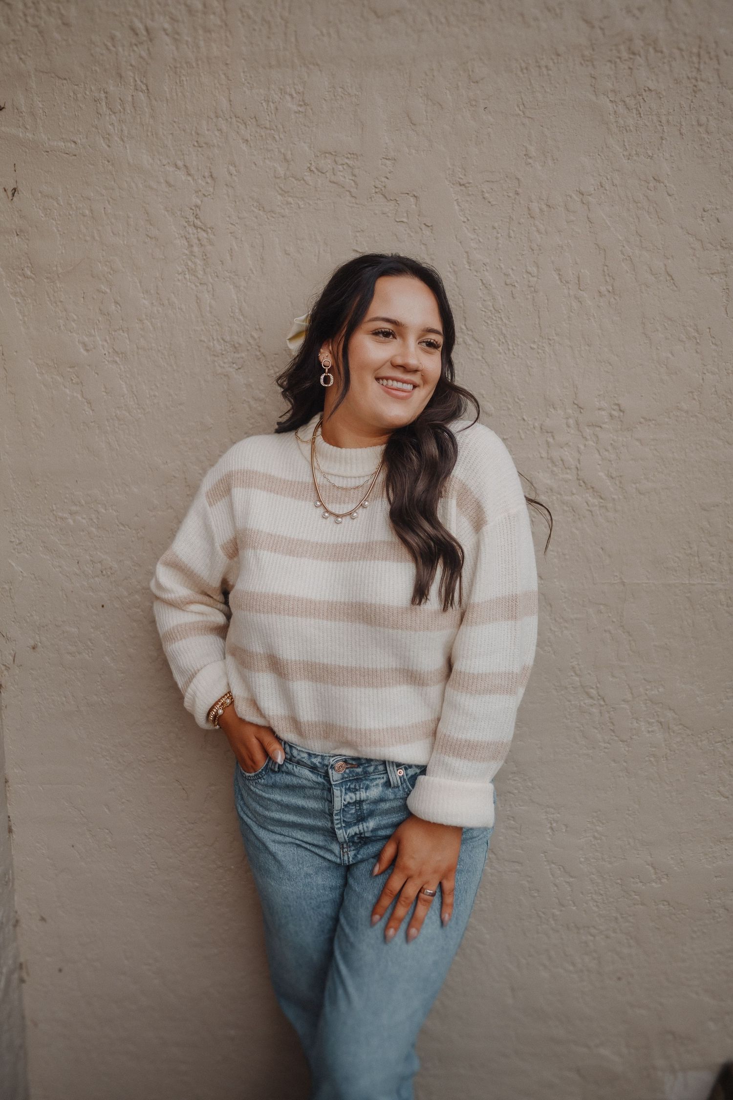 Woman in striped sweater and jeans poses casually against textured beige wall.