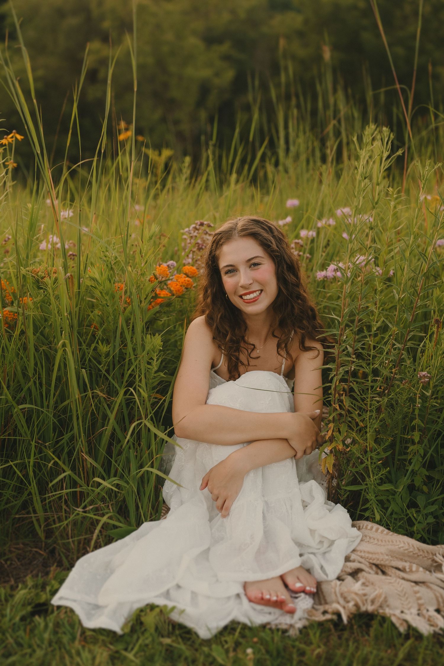 A casual portrait in tall grass shows someone seated on a blanket wearing a flowing white dress.