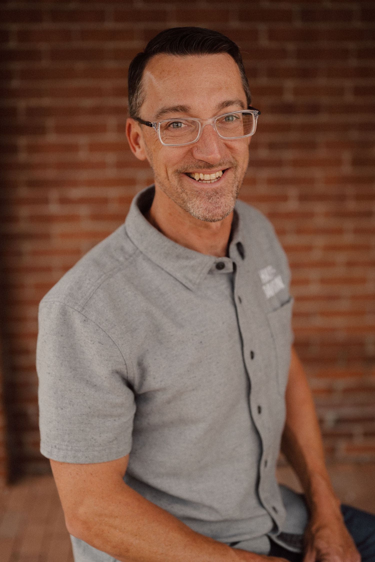 A person wearing glasses and a grey polo shirt smiles warmly against a brick wall background.