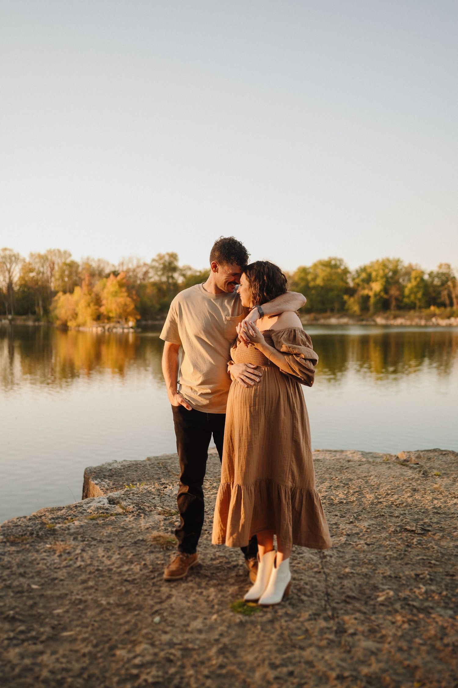 Sunset maternity photo of couple in casual attire embracing by lake.