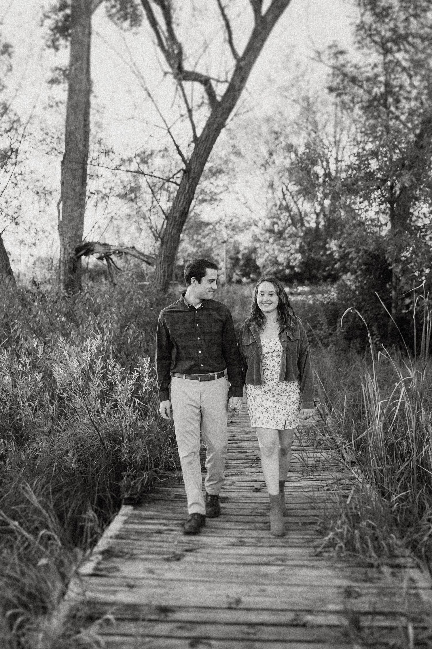 A couple takes a stroll together on a rustic wooden boardwalk path through tall grass.
