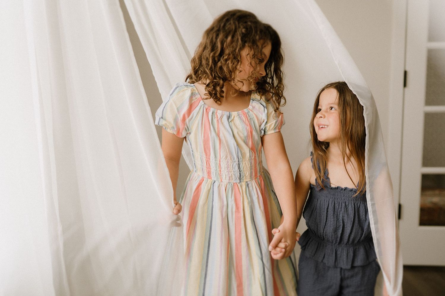 Two children in coordinating dresses holding hands against white backdrop.