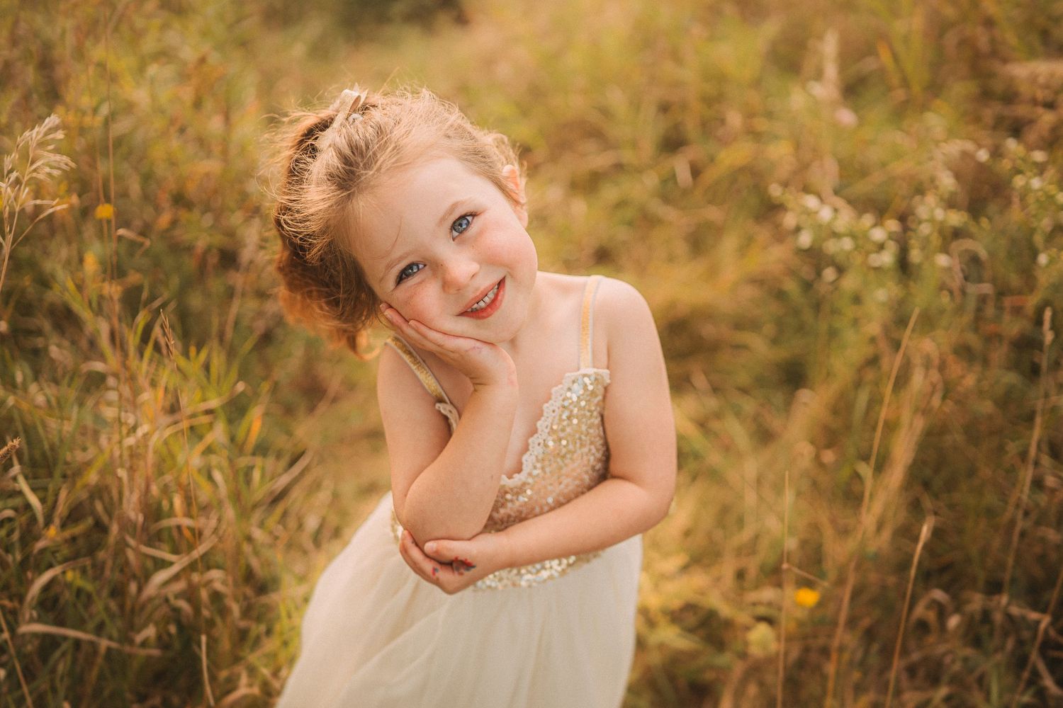 A child wearing a sparkly white dress poses playfully in a sunlit meadow.