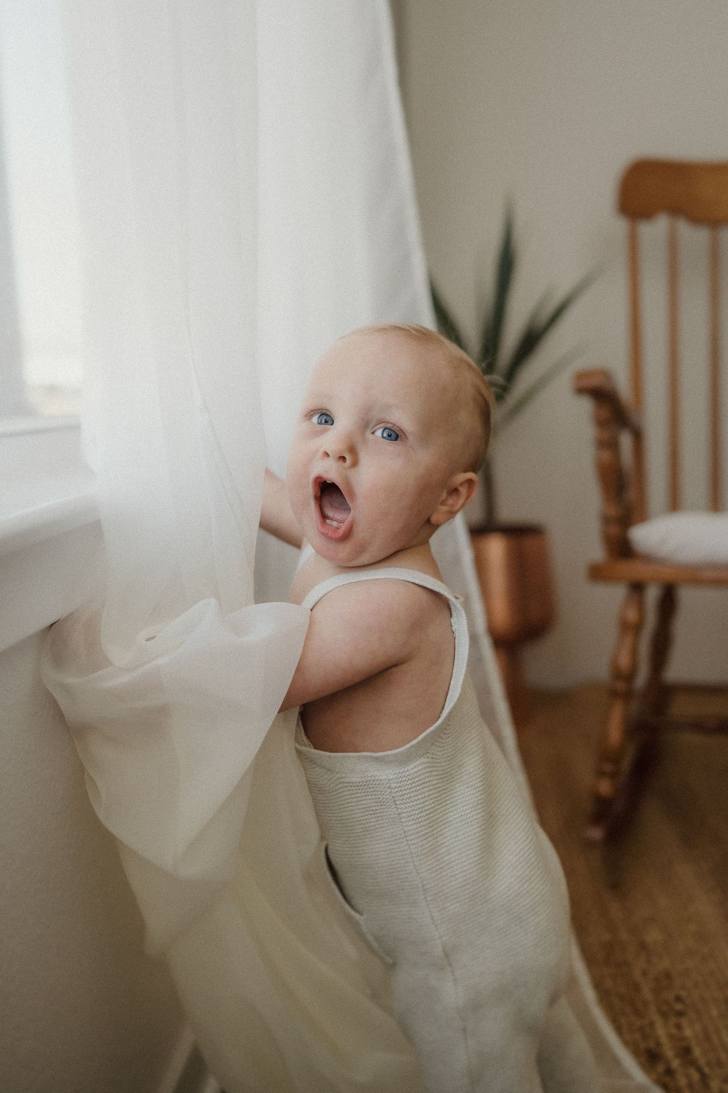 Baby in cream romper playing with white curtains near wooden rocking chair.