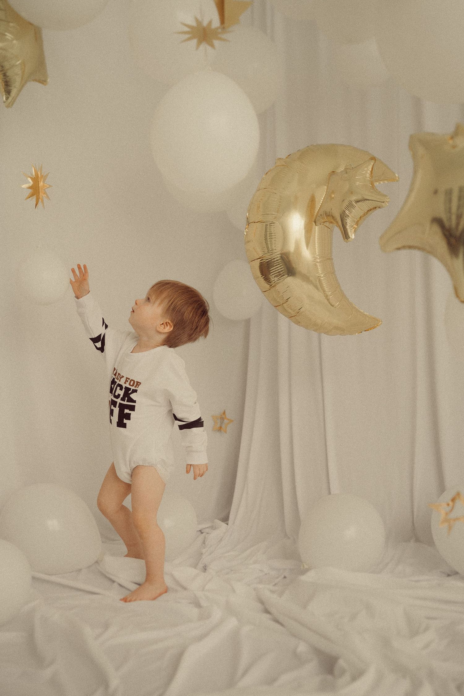 Child playing with gold star and moon balloons against white backdrop in sports themed onesie.