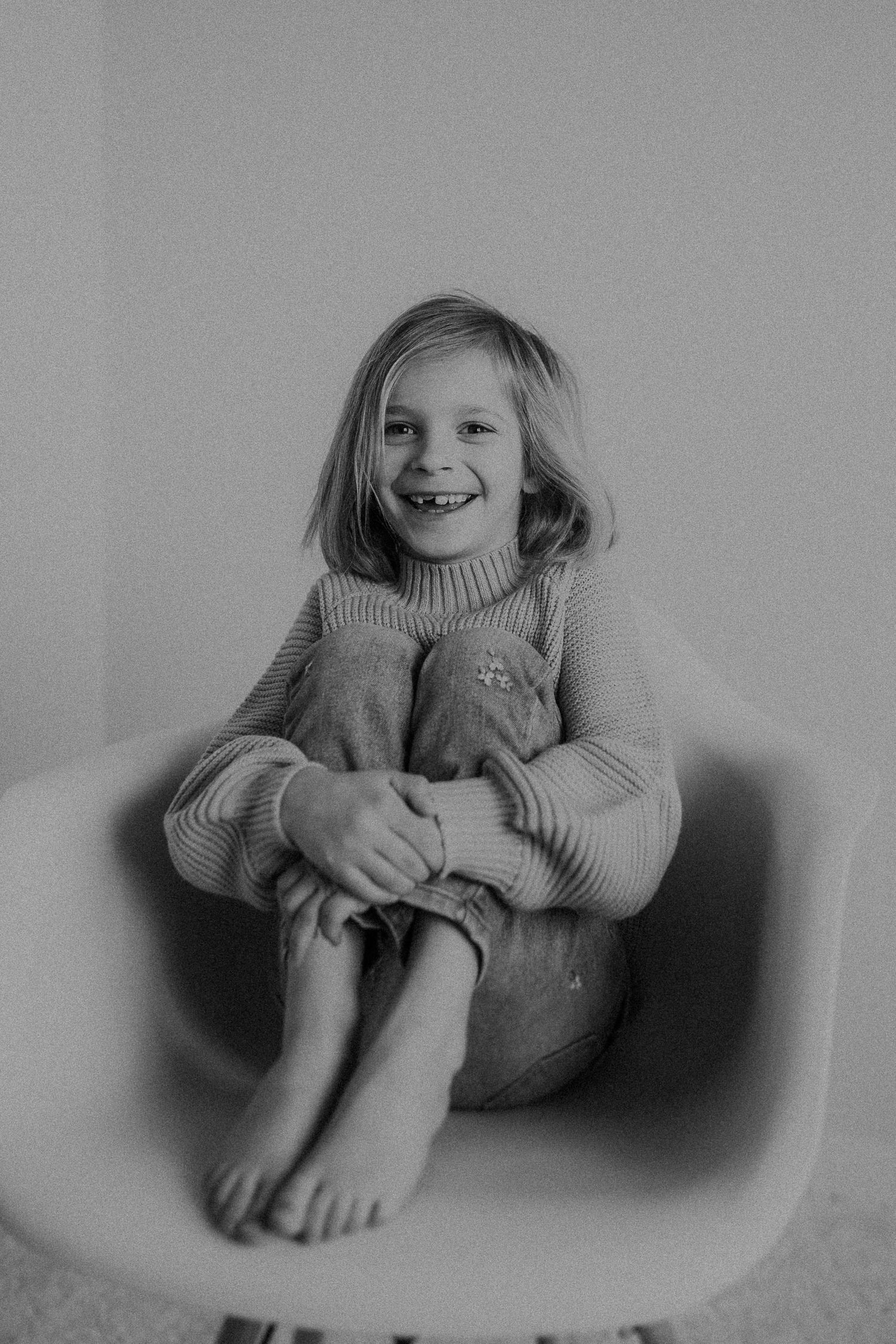 Joyful child in sweater sitting comfortably on modern chair in monochrome photo.