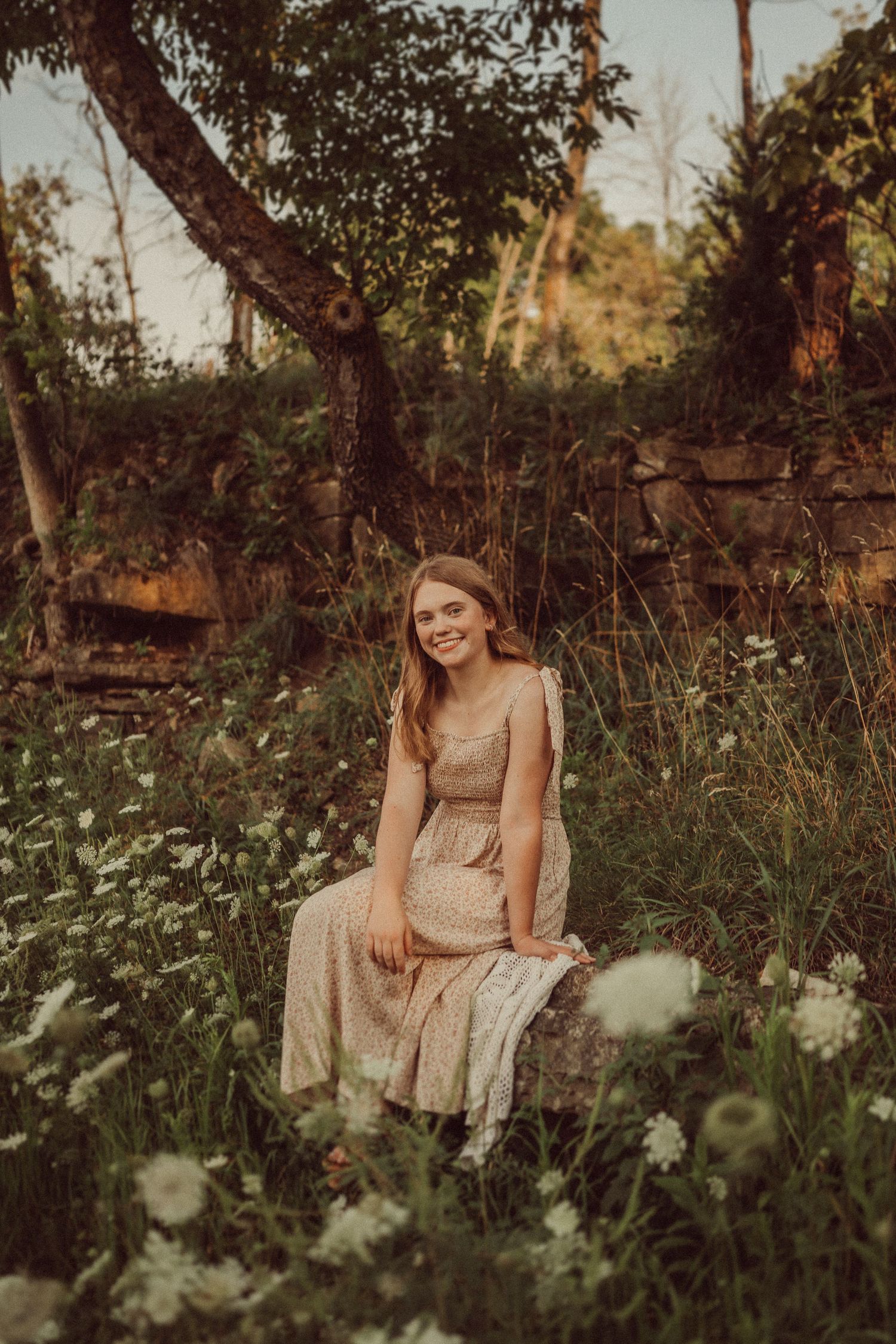 A romantic outdoor portrait with stone wall backdrop and delicate wildflowers in warm sunset lighting.