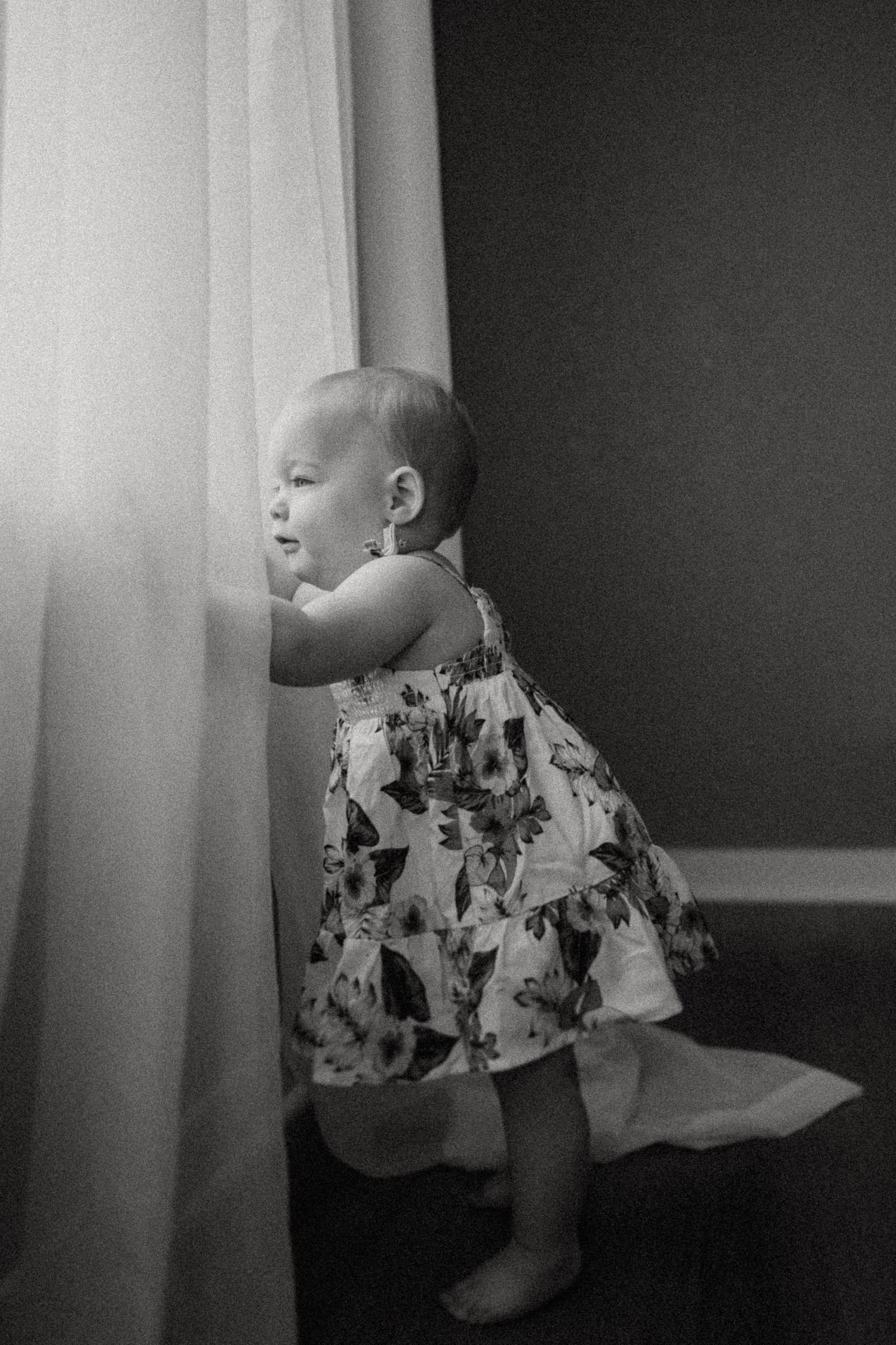 A baby in a floral dress peeks out from behind a white curtain in a black and white photograph.