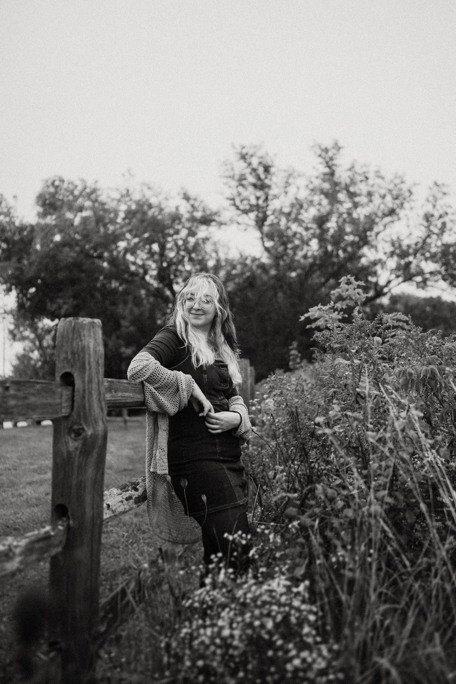 Candid black and white portrait by rustic wooden fence surrounded by wildflowers.