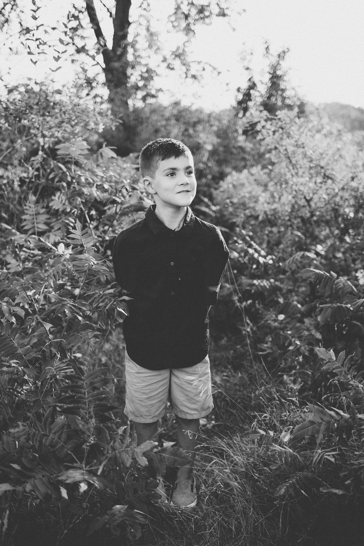 Black and white portrait of child standing in tall grass and foliage outdoors.