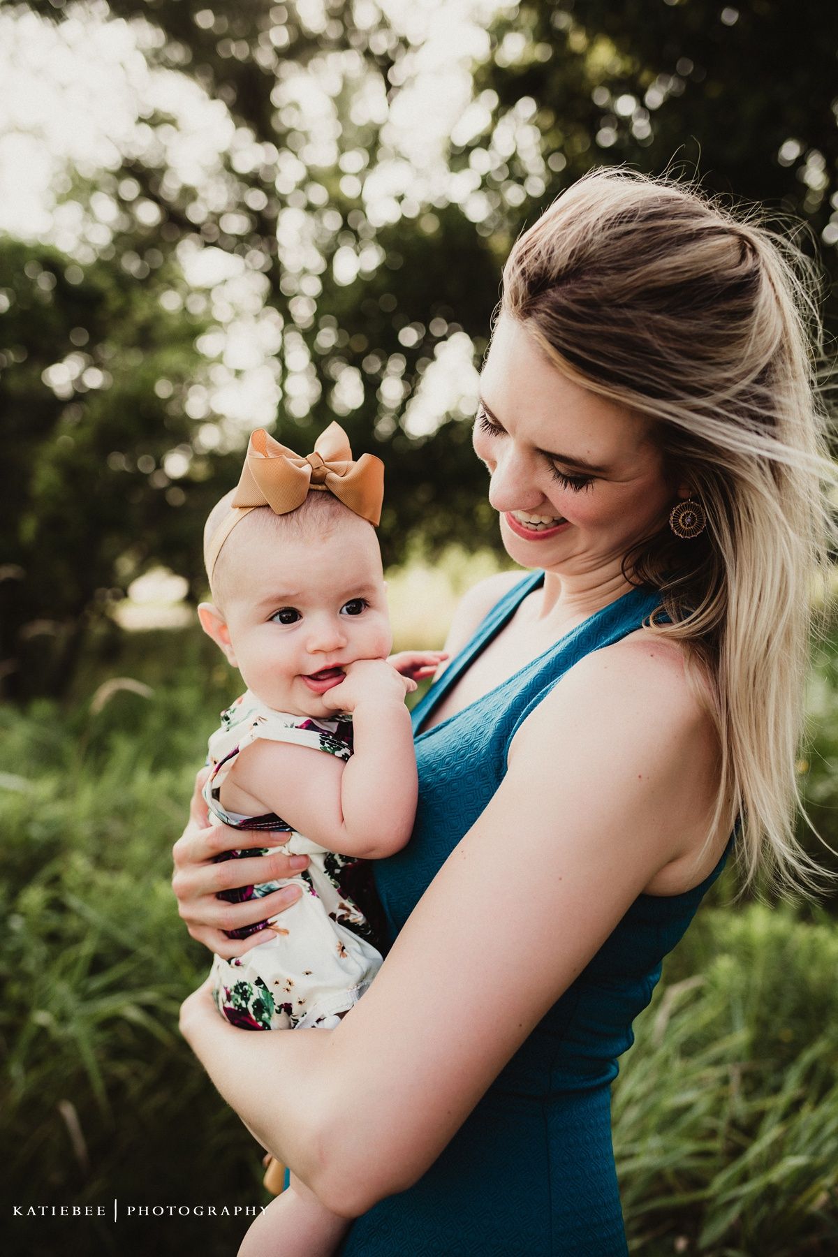 Parent in teal tank top holds smiling baby with tan bow against natural outdoor background.