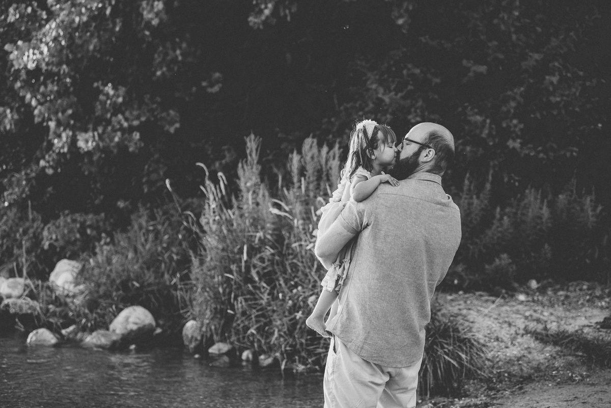 Black and white portrait of person lifting small child by lakeside vegetation.