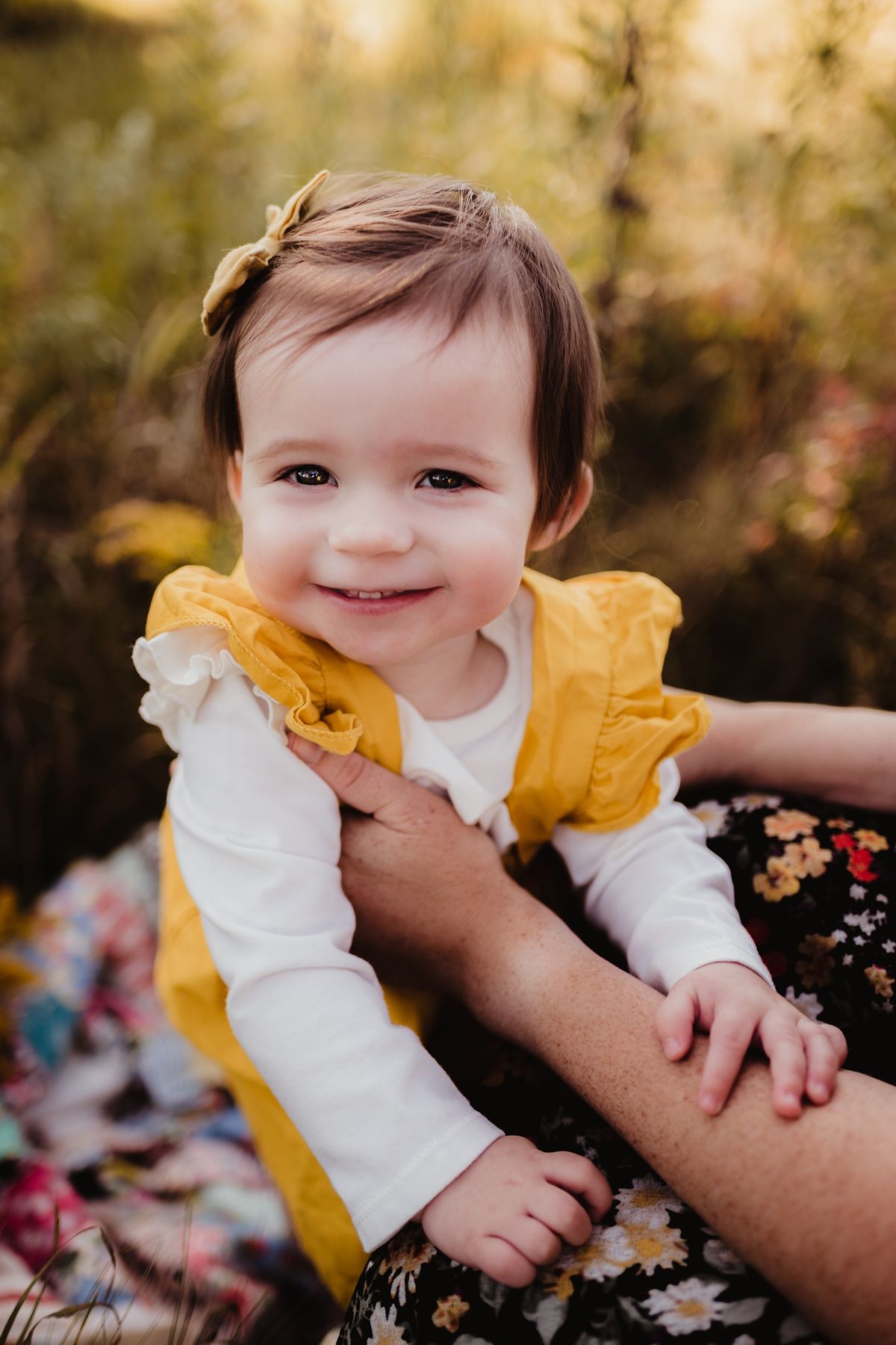 A toddler in a yellow dress smiles while sitting outdoors against an autumn background.