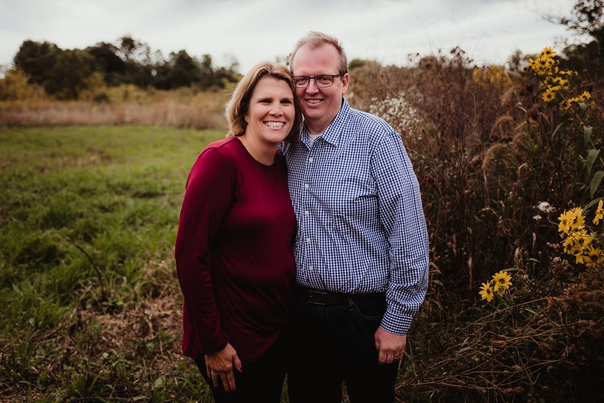 A couple wearing burgundy and blue smiles together against an autumn landscape.