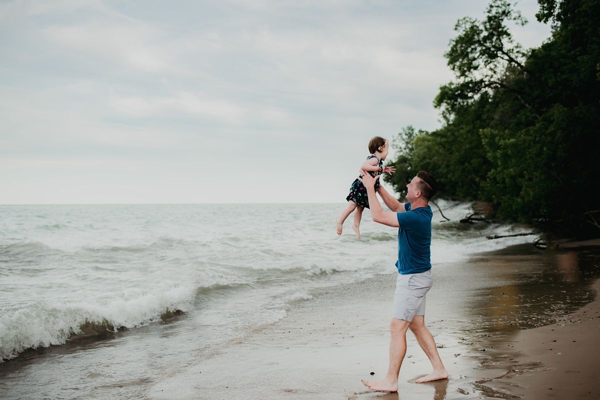 Parent lifting child playfully by the ocean shore on a cloudy day.
