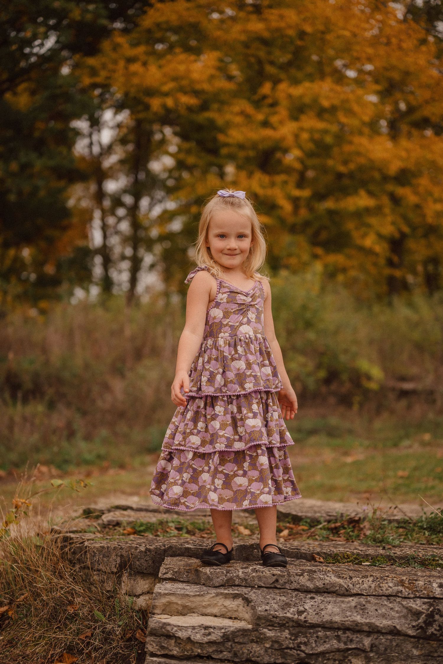 Child in a tiered floral dress standing on stone steps with autumn foliage behind.