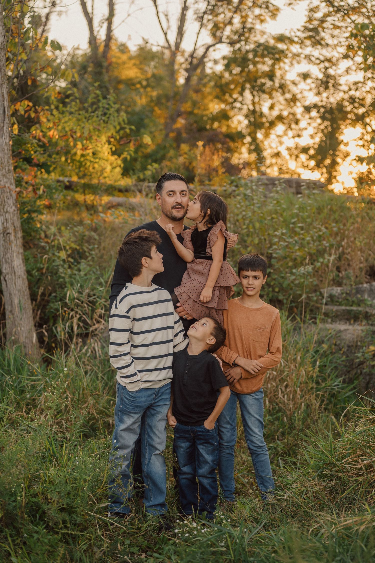 A family group poses together outdoors during golden hour in an autumn nature setting with trees and tall grass.
