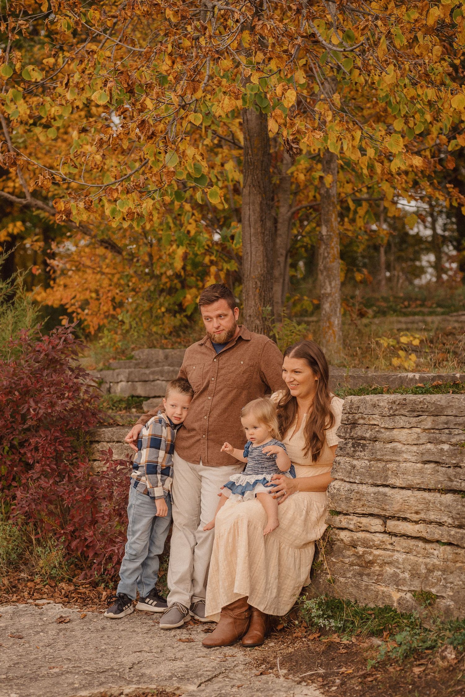 A family sits together on a stone wall surrounded by vibrant fall foliage in a scenic autumn setting.