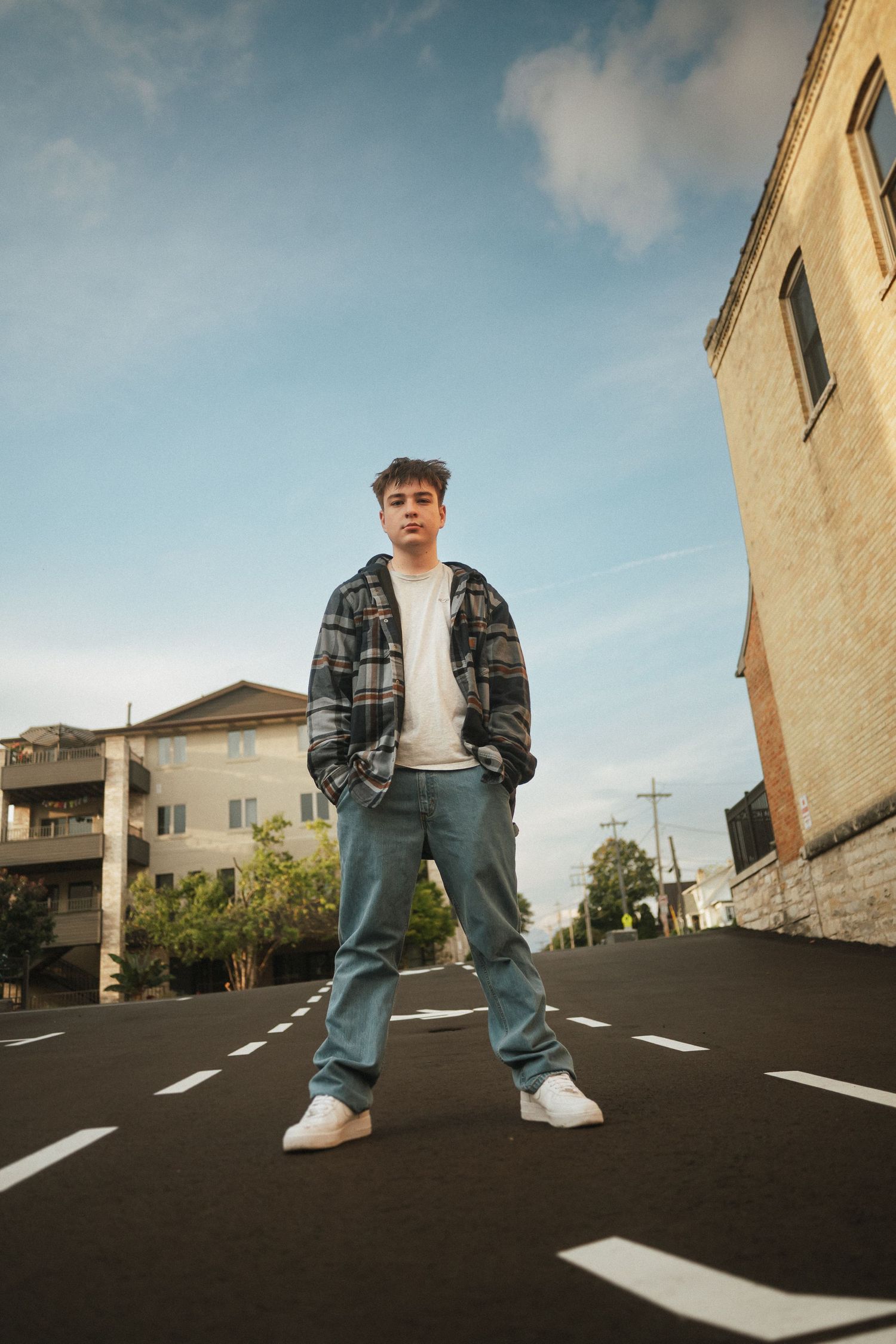 Wide angle shot of person in plaid jacket posing on parking lot with urban buildings in background.