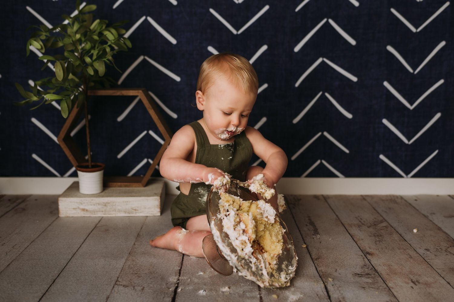 Baby in olive romper examines smashed cake on wooden floor during photo session.