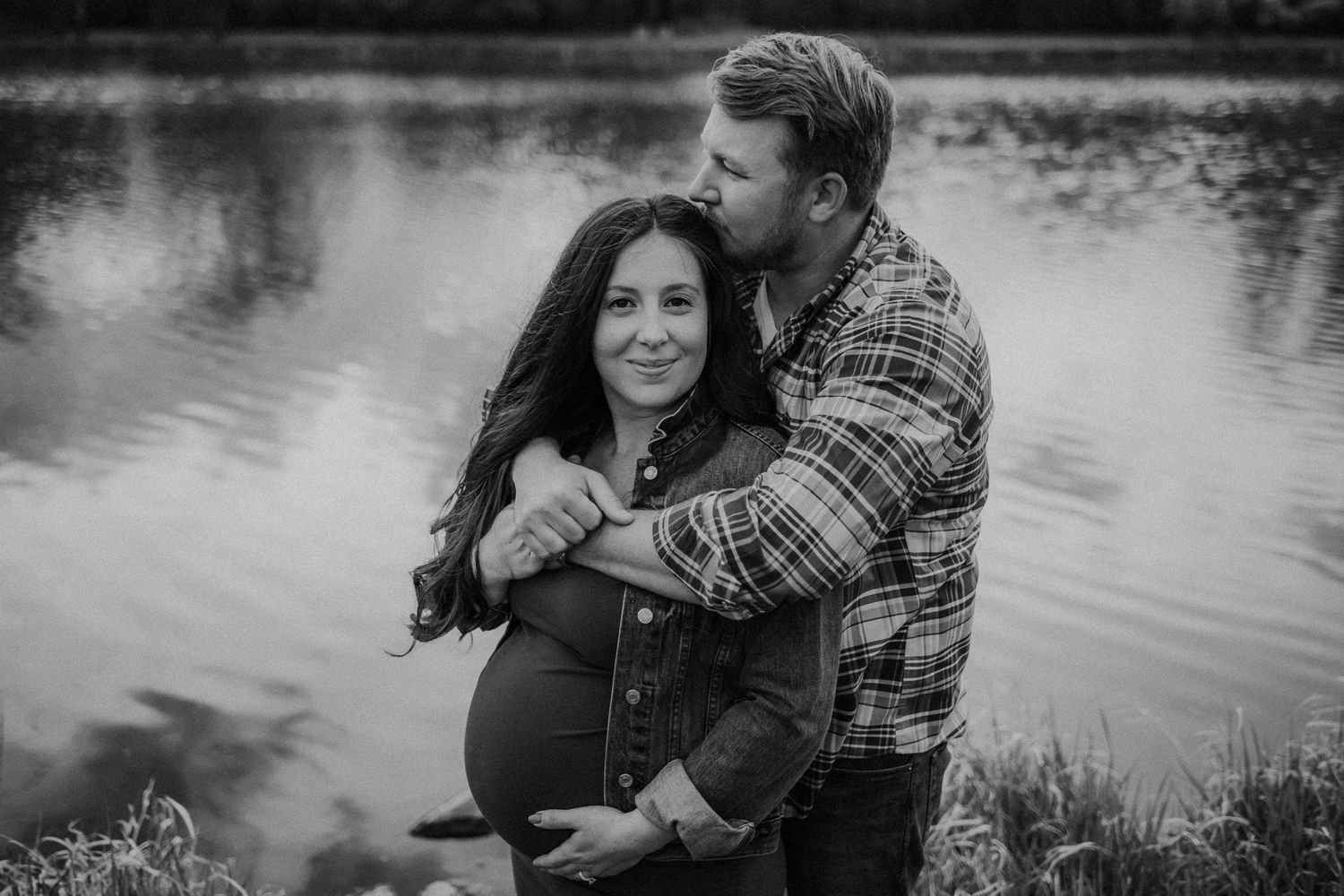 Black and white maternity portrait of couple embracing by serene lakeside.