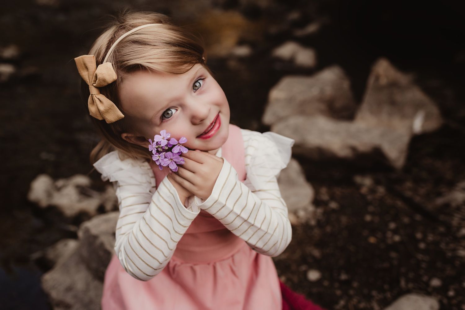 A child holds purple flowers while wearing a pink dress and cream sweater.