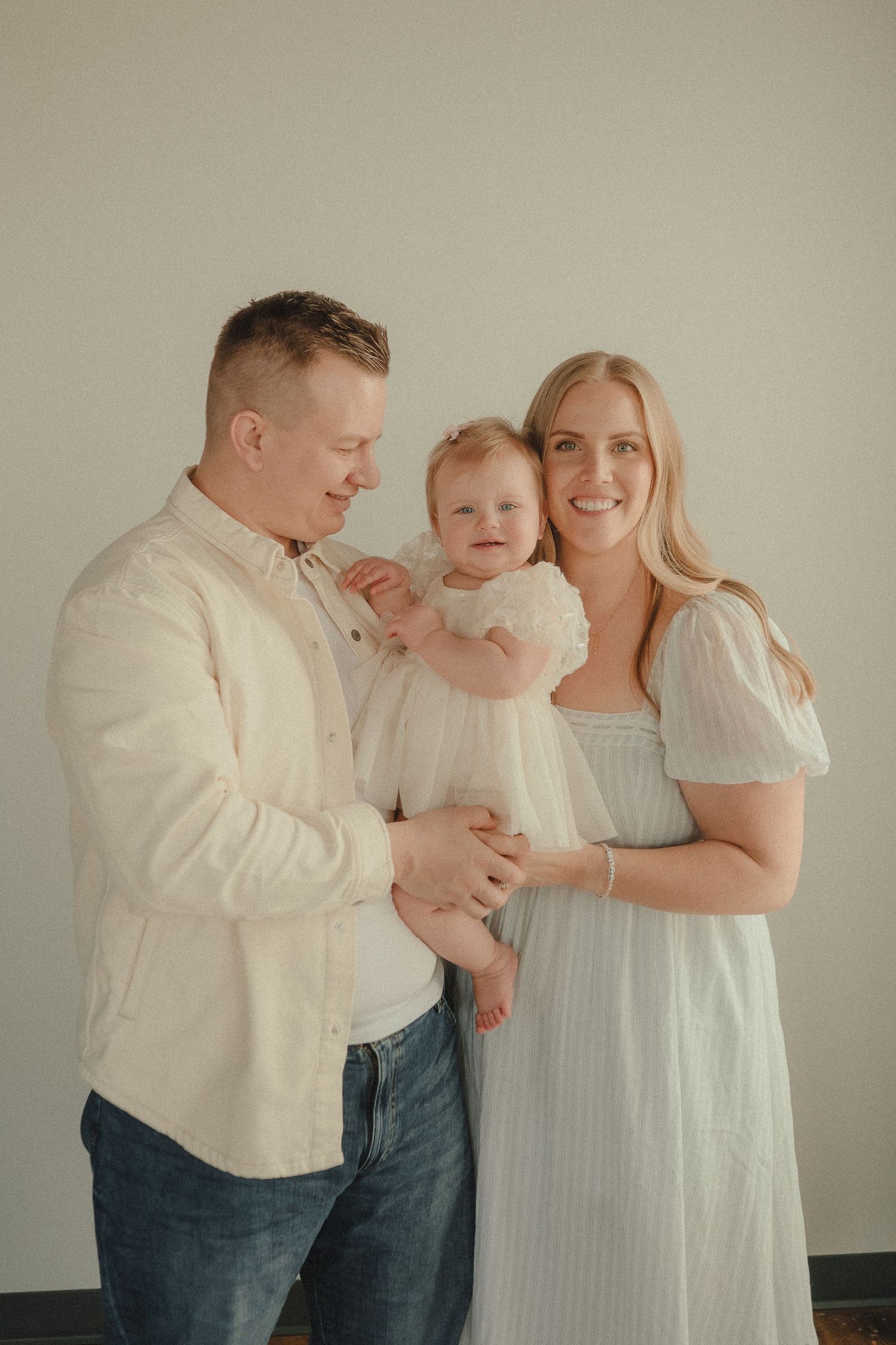 A smiling family of three poses together in a bright studio, parents holding their happy baby girl in white.