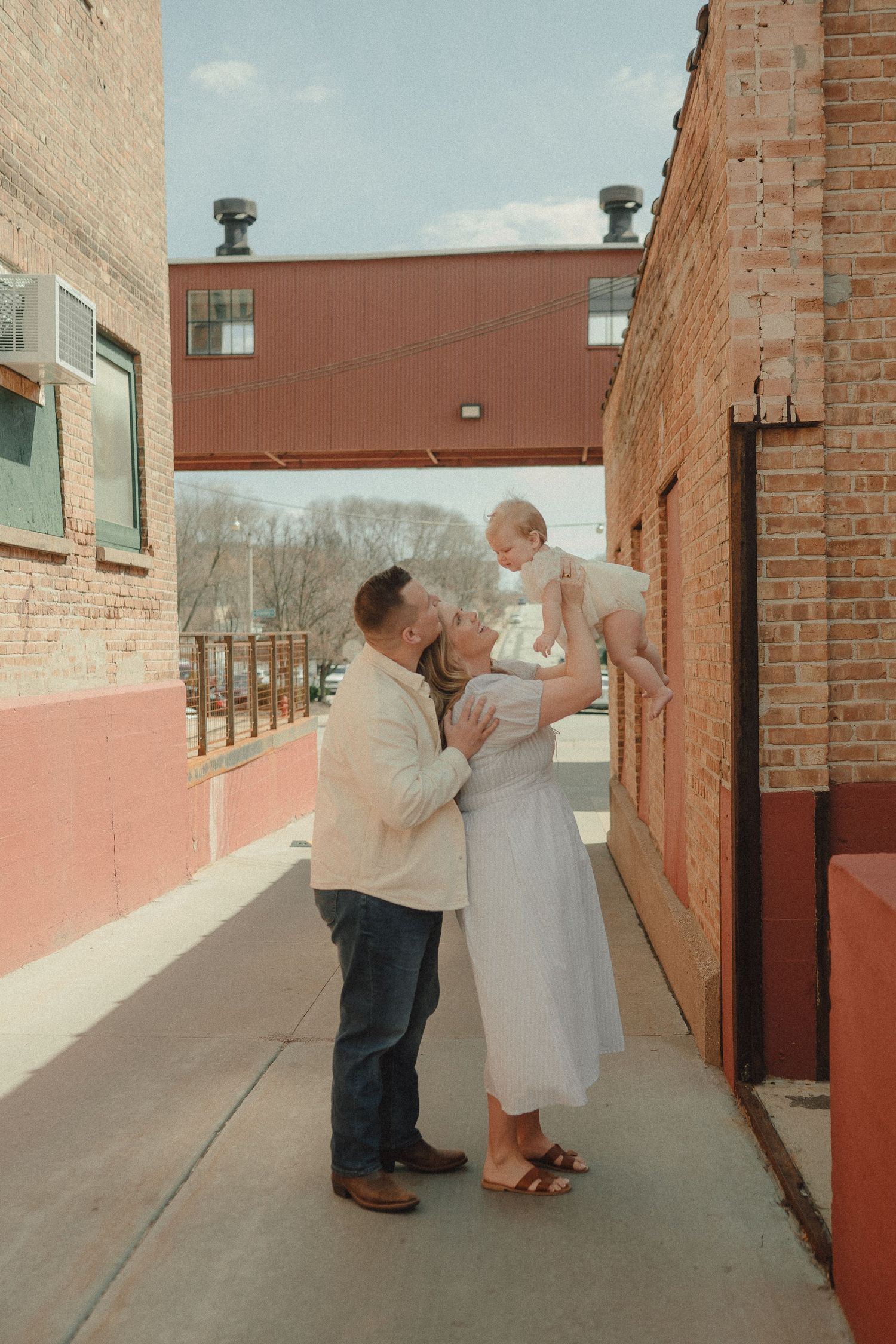 A couple shares a kiss while lifting their baby girl in a charming brick alleyway during a family photo session.