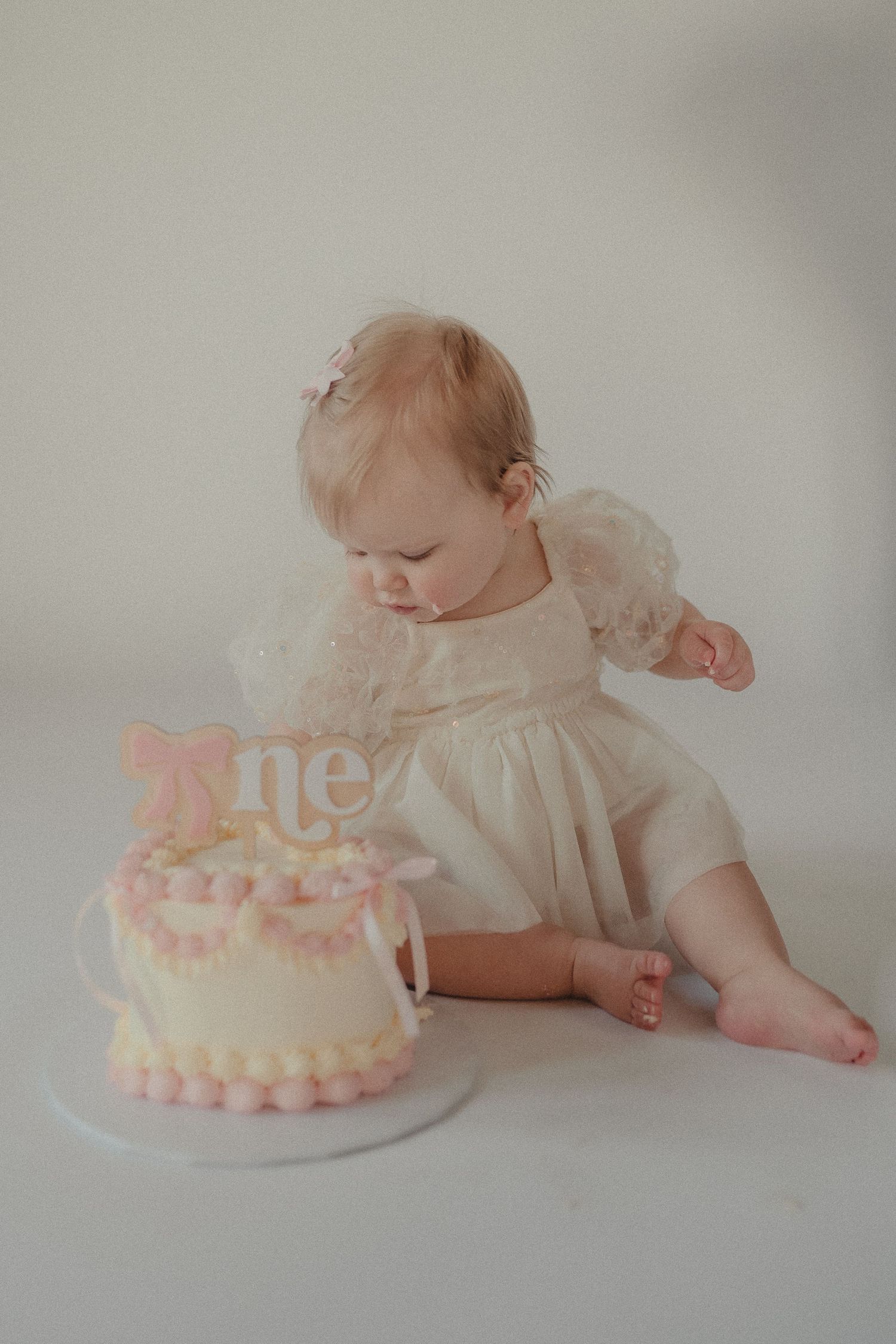 Baby girl in white lace dress examines her pink and white smash cake with a one topper at her first birthday.