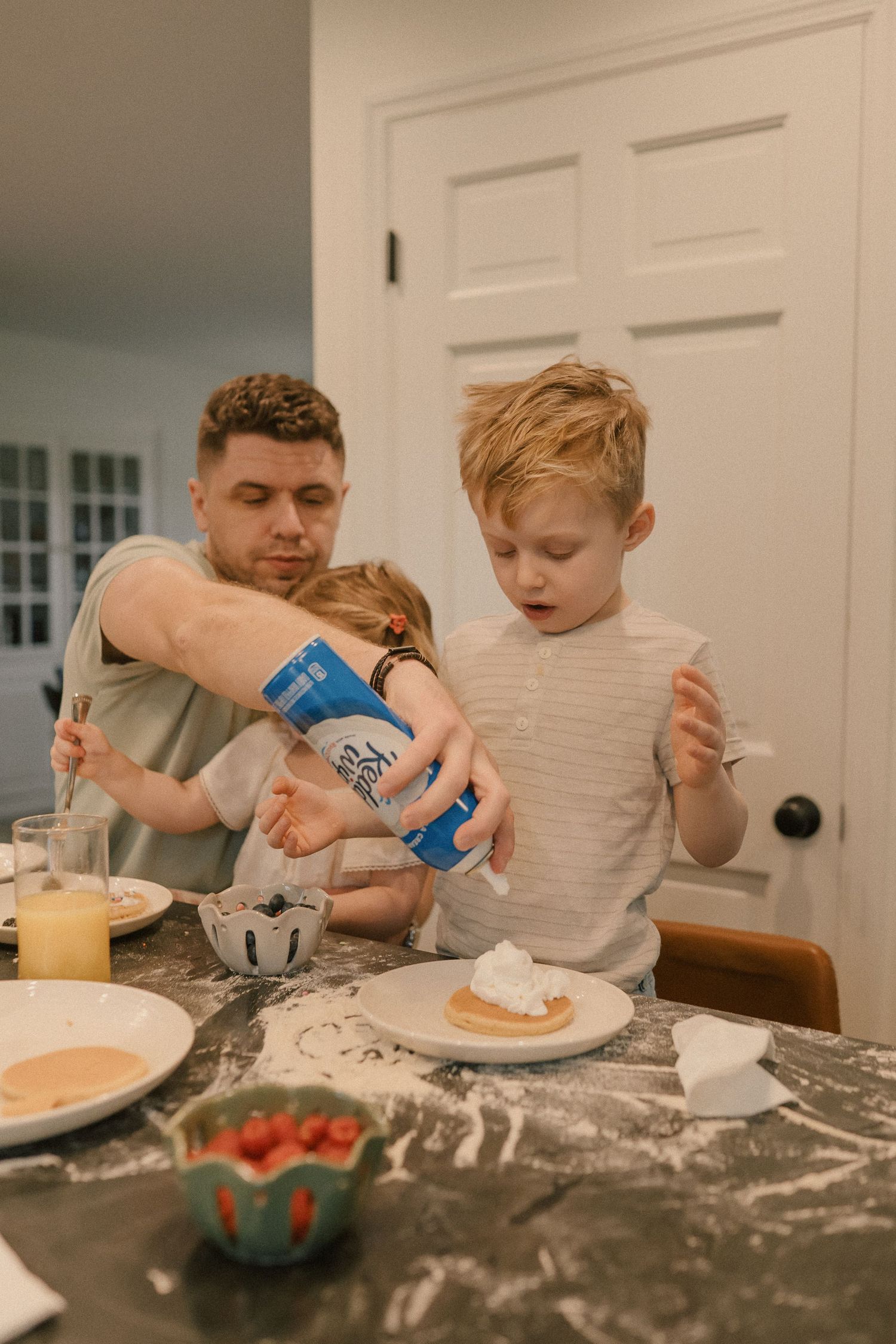 Father sprays whipped cream on pancakes while two young children watch at a messy kitchen table.