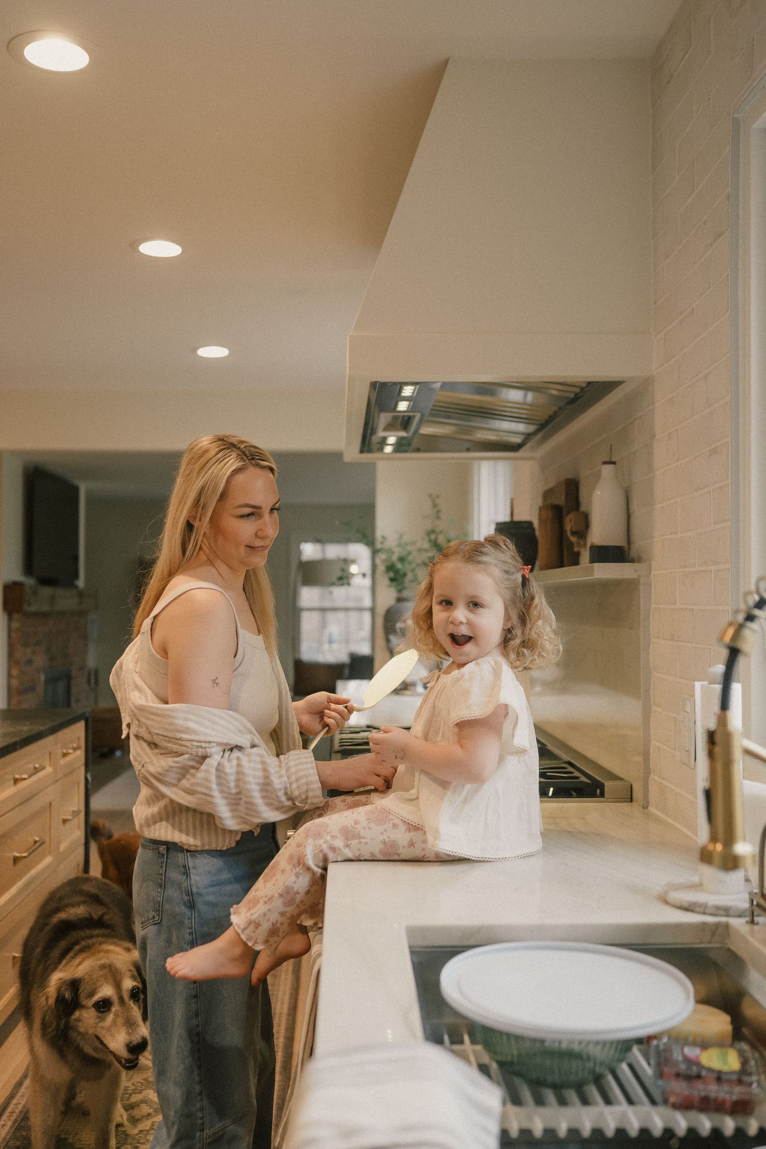 A smiling mother holds her laughing toddler daughter on a kitchen counter while a dog watches.