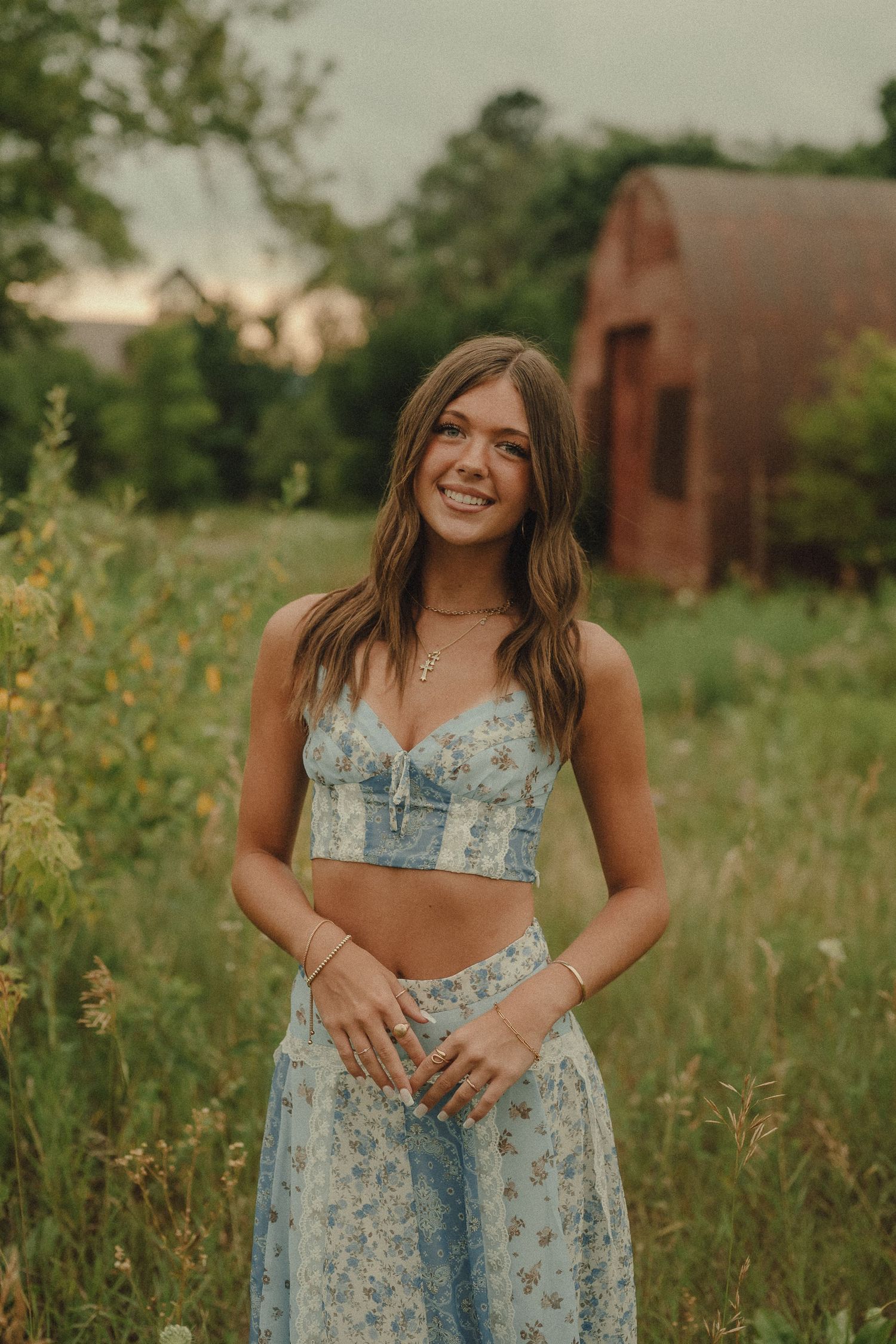 Smiling young woman in blue floral crop top and skirt posing in wildflower field at sunset.