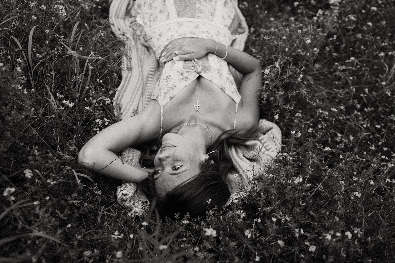 Black and white photo of a smiling woman lying in a wildflower meadow, wearing a floral dress.