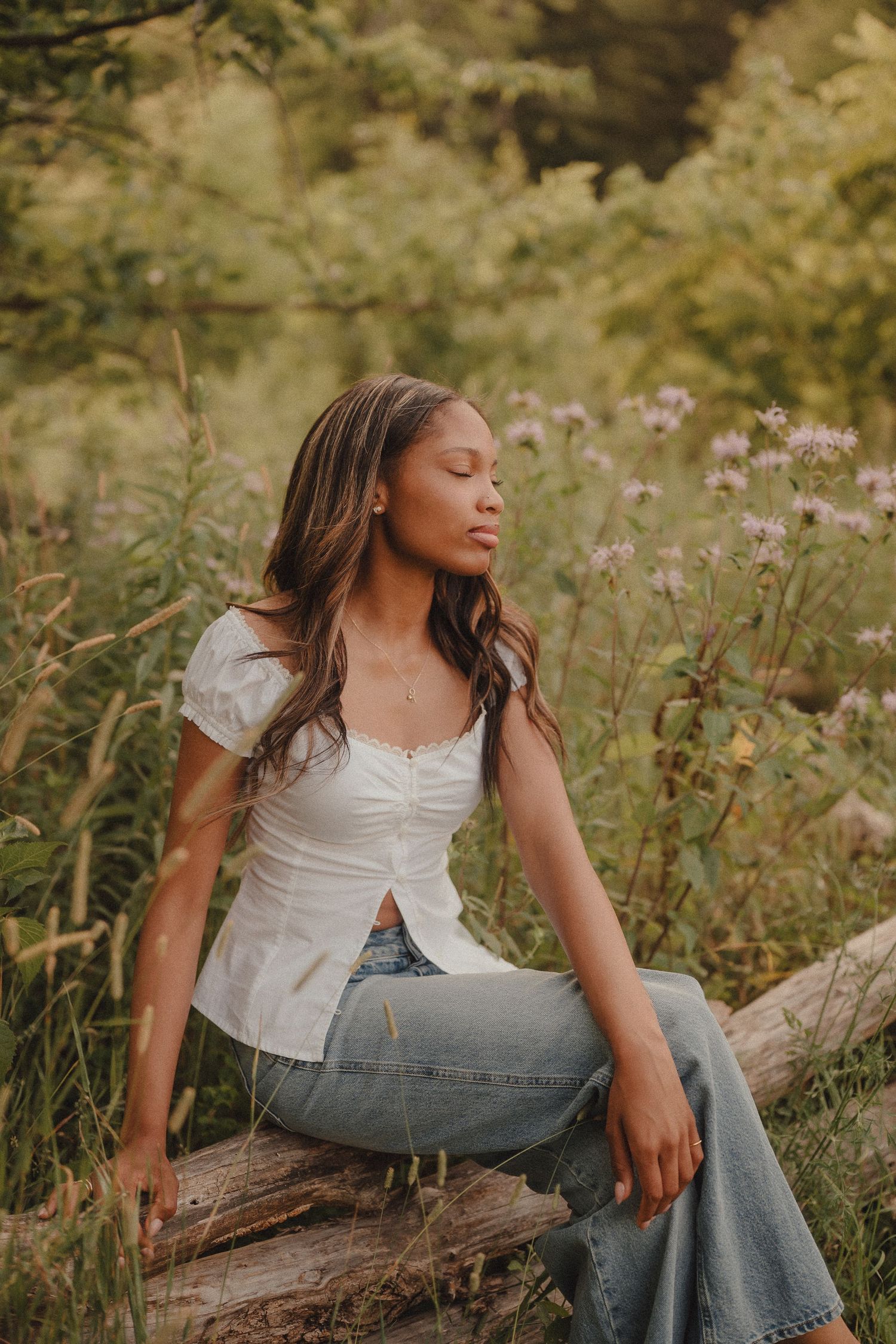 Young woman sitting on a log with eyes closed among wildflowers, wearing white top and jeans.