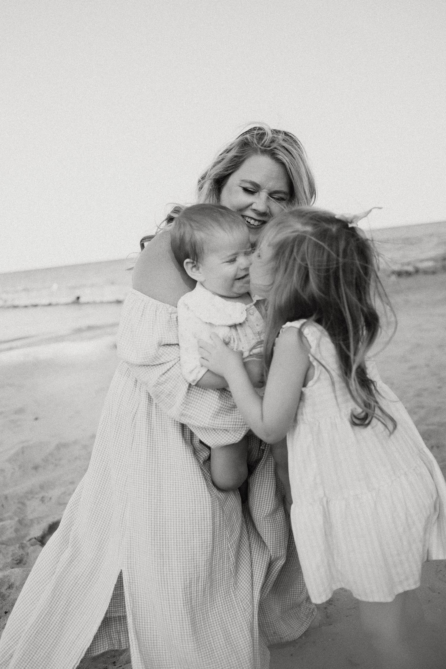 Black and white photo of a smiling mother holding a baby while an older daughter kisses the infant on a beach.