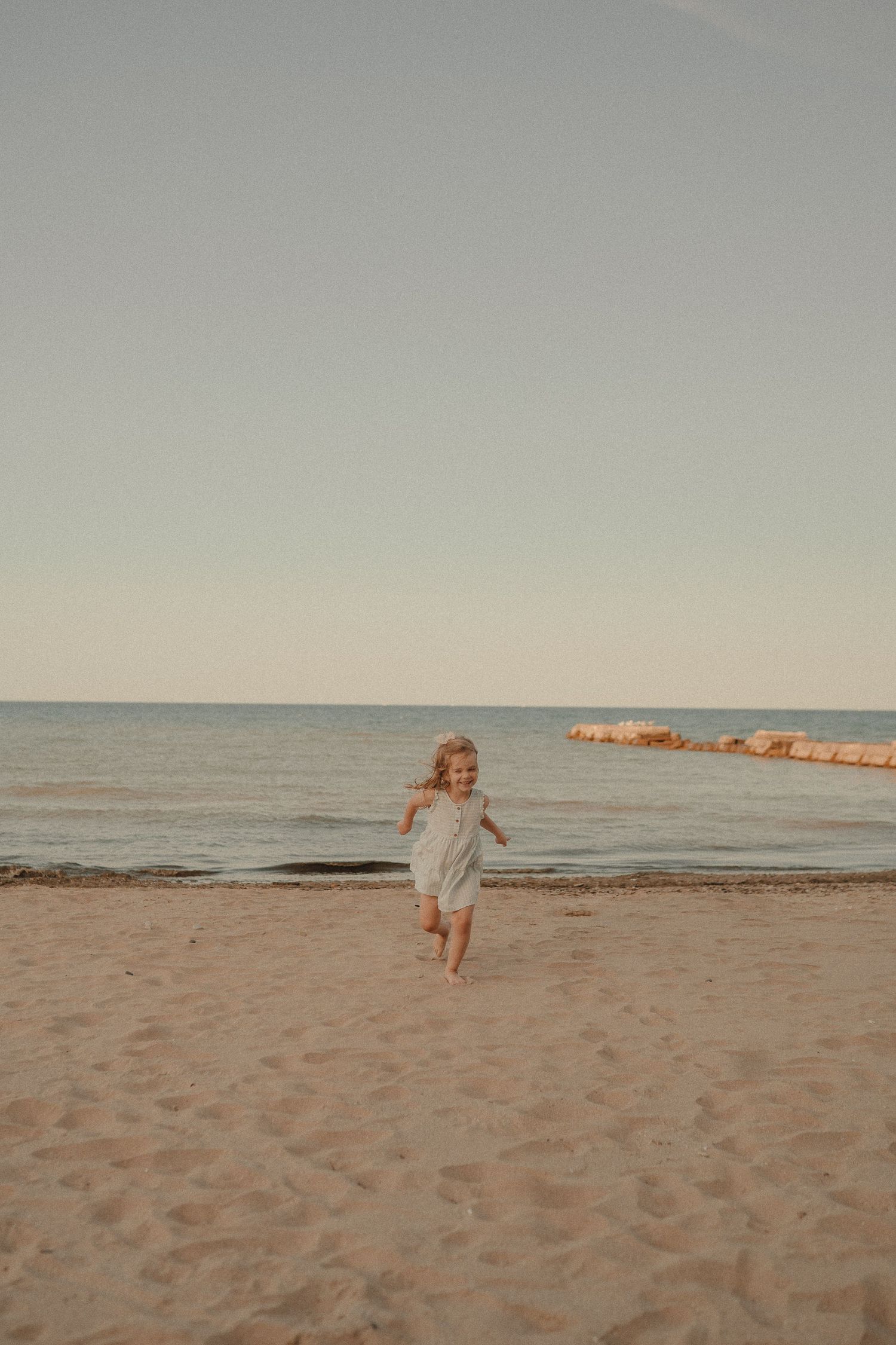A happy young girl in a light blue dress runs barefoot along a sandy beach with calm ocean waters behind her.