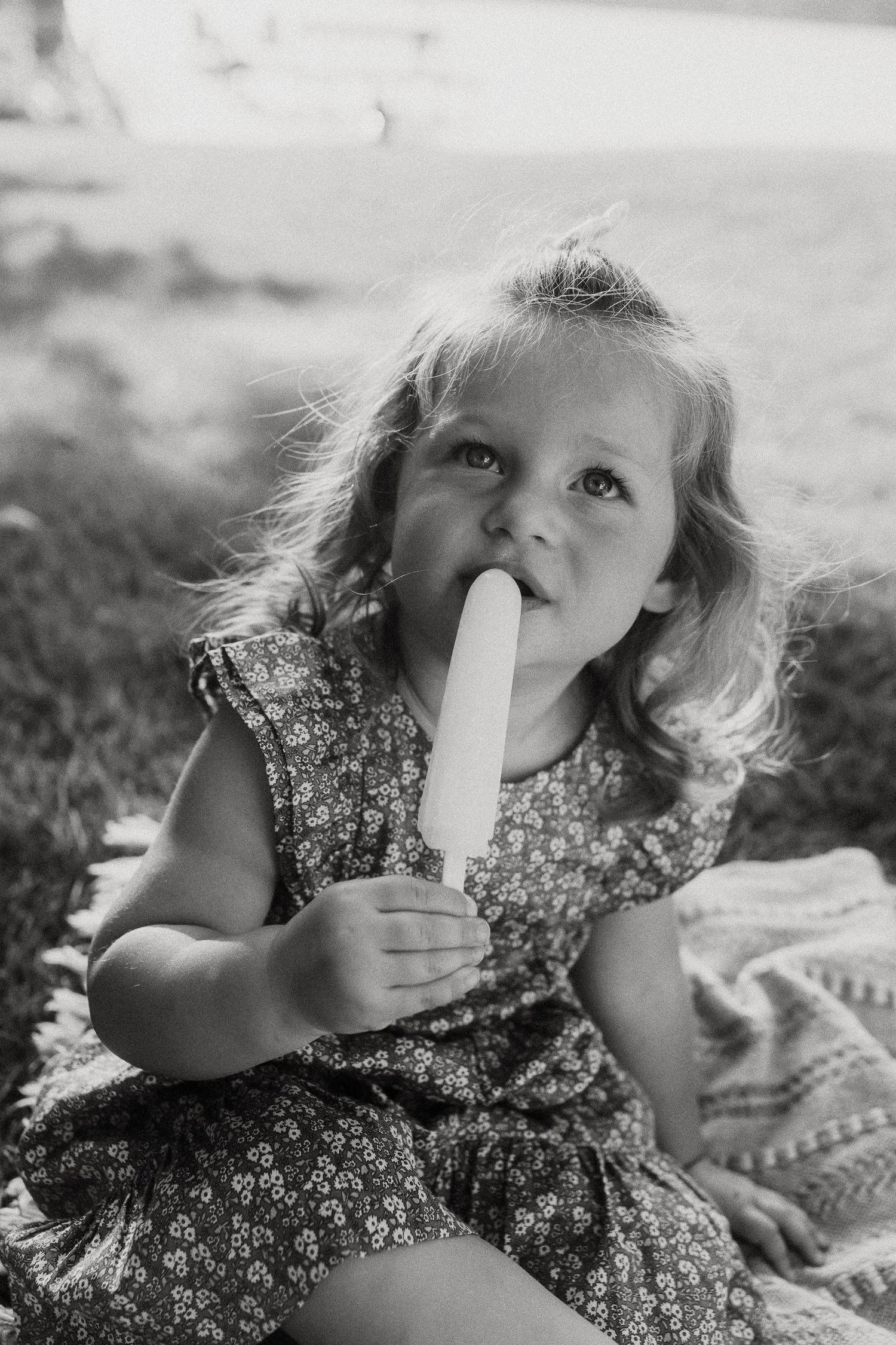 Black and white portrait of a curly-haired toddler girl eating a popsicle while sitting on grass.