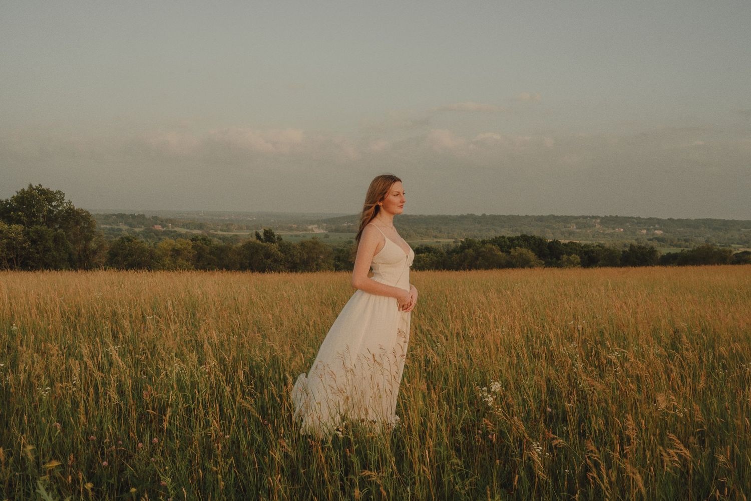 A bride in a white lace gown stands gracefully in a golden meadow at sunset with rolling hills in the background.