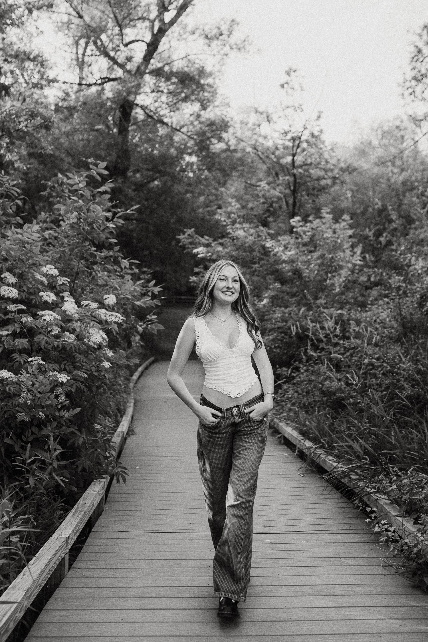 Young woman in jeans and white top walks along a wooden boardwalk surrounded by lush green foliage.