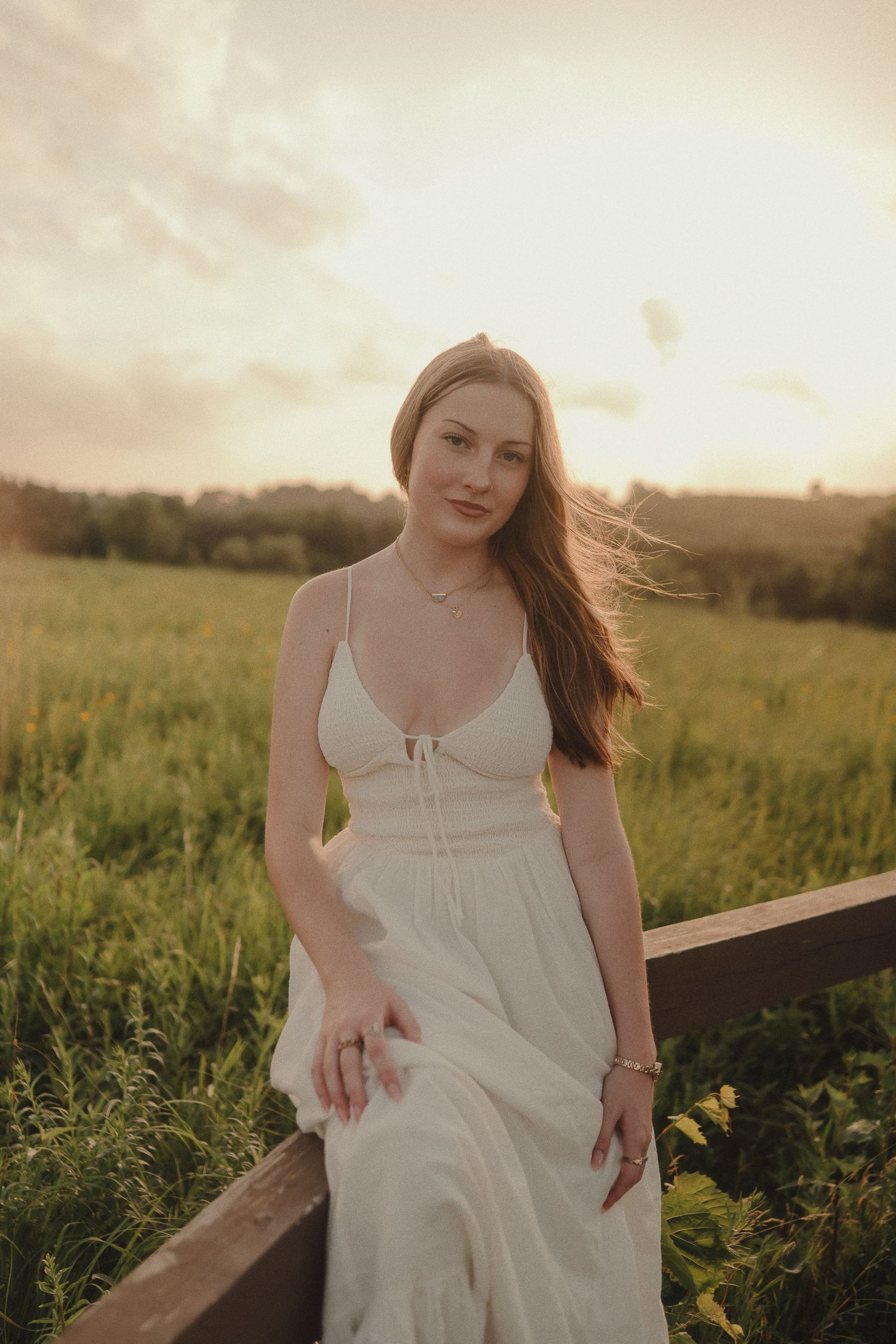 Young woman in white sundress poses by wooden fence in golden sunset light with green meadow background.