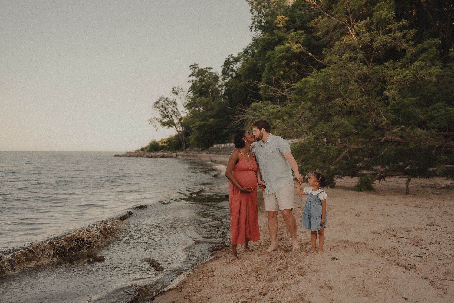 Pregnant couple with child on sandy beach near water, sharing a tender kiss at golden hour with lush trees behind.