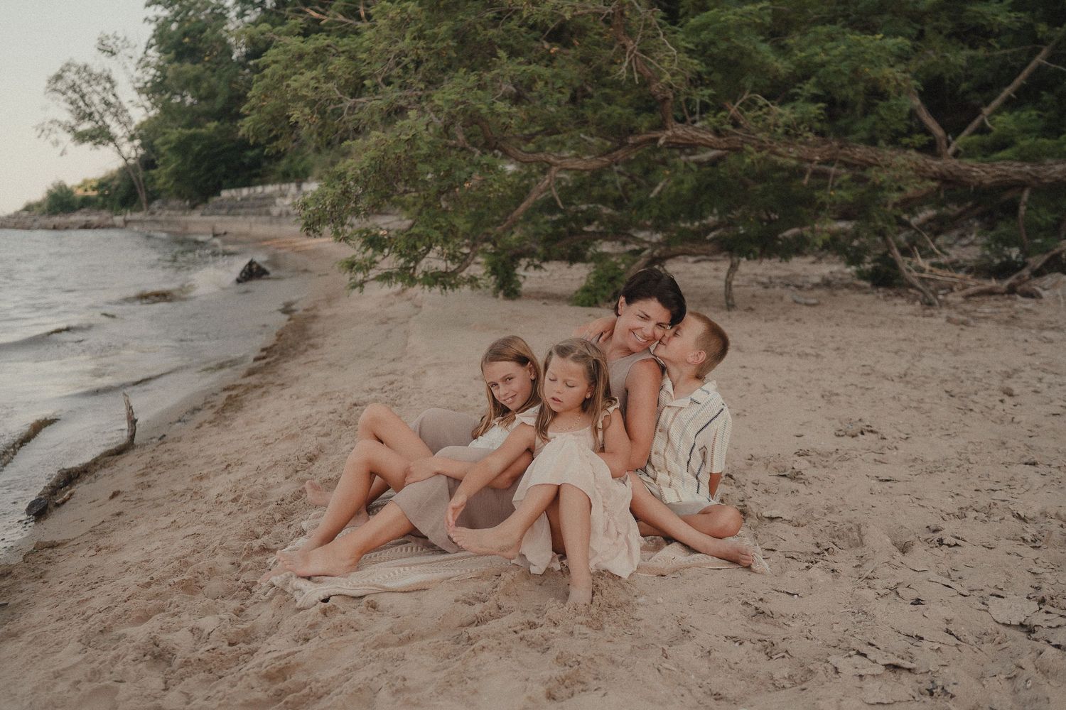A mother sits on a sandy beach hugging her three children under shaded trees near the water.