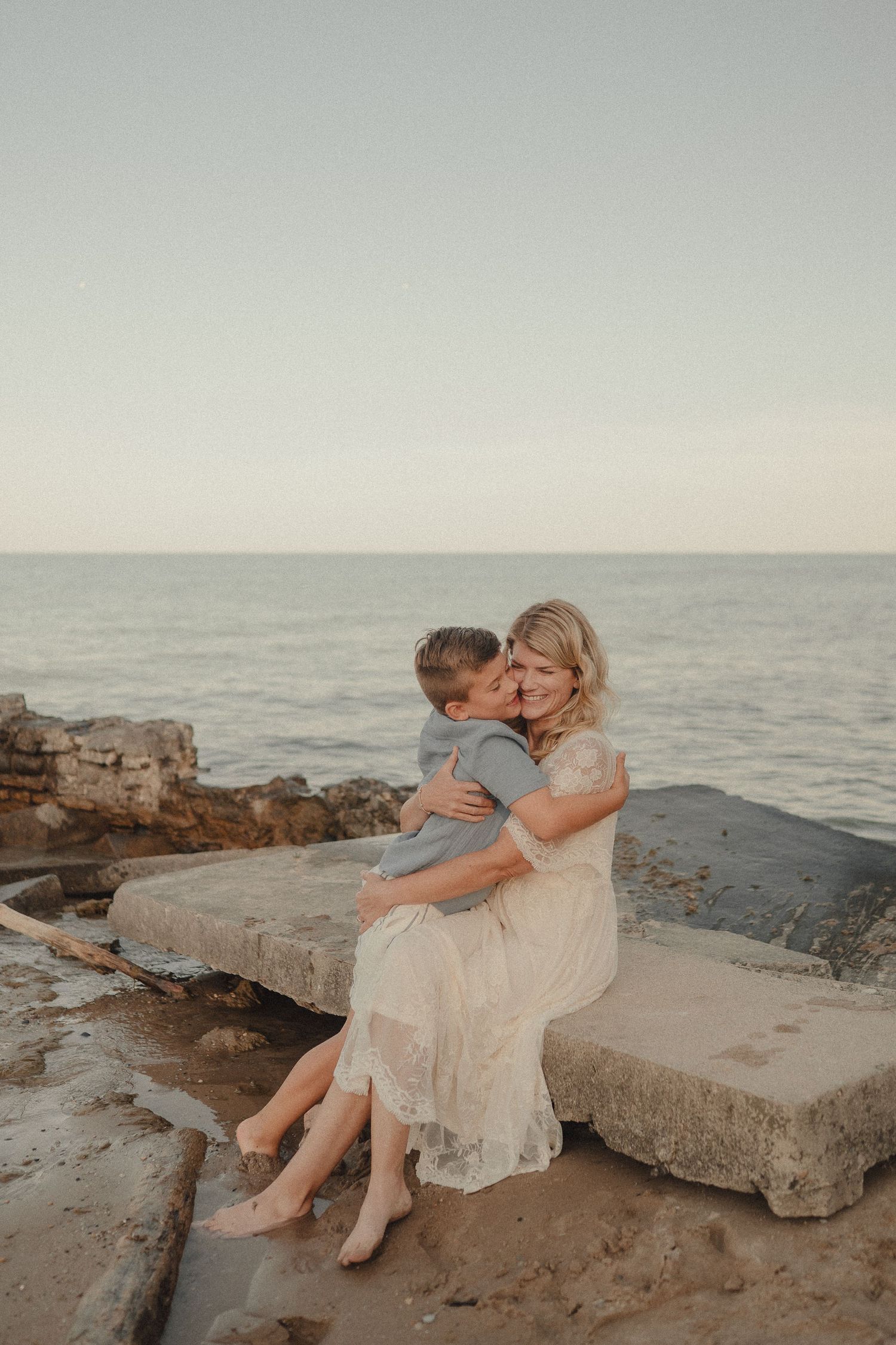 Mother and young son share a tender embrace while sitting on rocky seaside steps during a golden hour beach session.